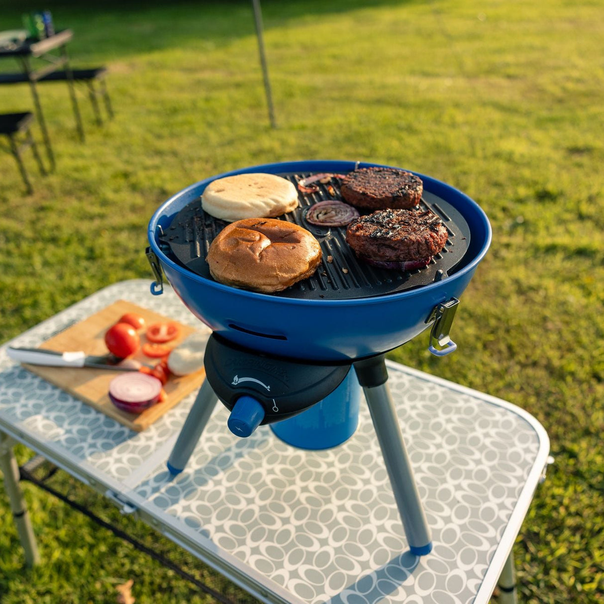 kompakts grils ar burgeriem stāv uz piknika galda